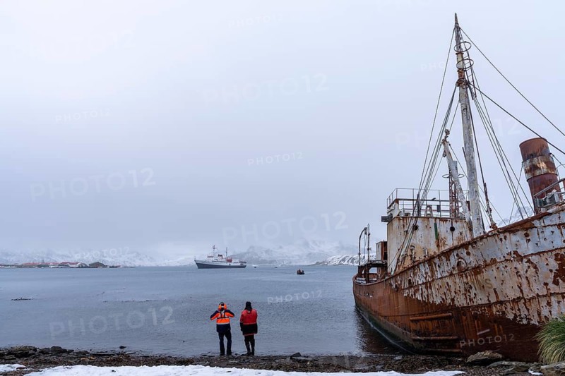Rusted wreck of the whaling ship Petrel on the shore, two people in expedition clothes looking at the bay with an expedition ship and zodiac, snow-covered mountains in the background, former Grytviken whaling station, South Georgia