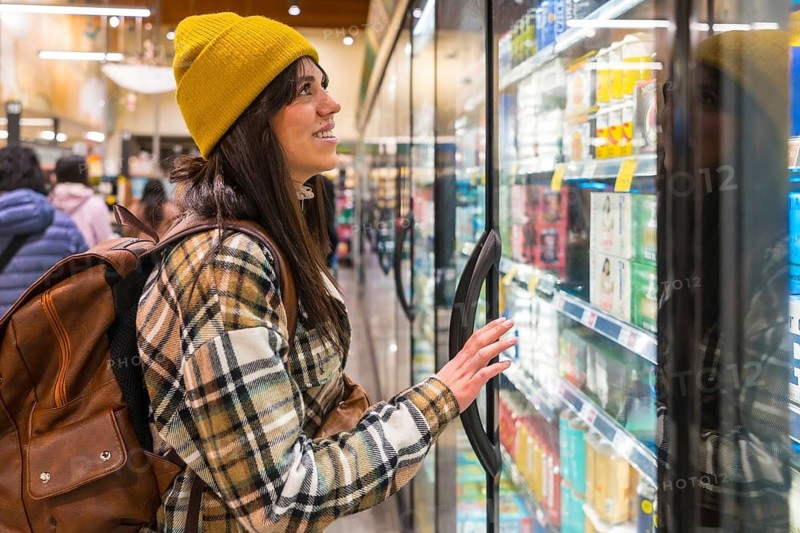 Young woman wearing a yellow beanie and plaid shirt browsing refrigerated beverage aisle in a grocery store, making a purchase decision while looking at various products
