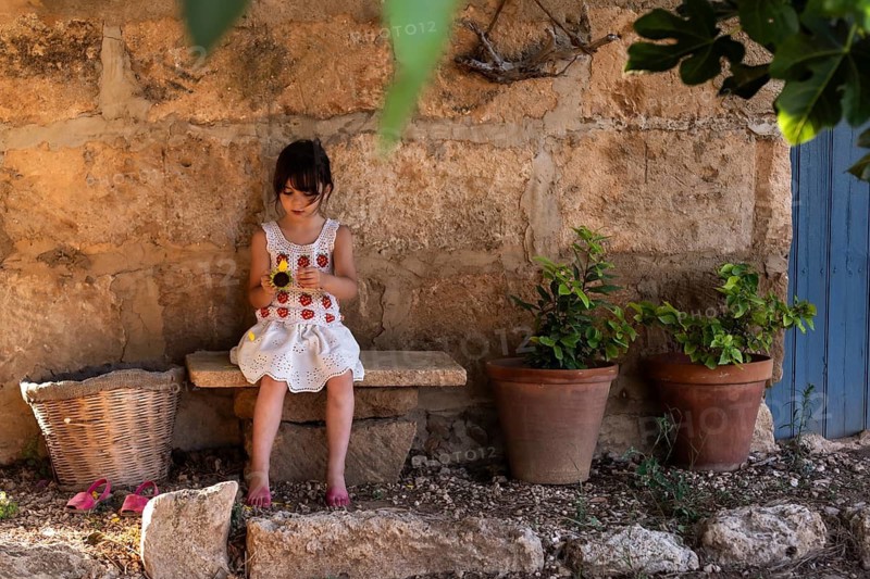 A young girl in a white dress sits on a stone bench, holding a sunflower. She is surrounded by potted plants against a rustic stone wall. Her sandals rest nearby