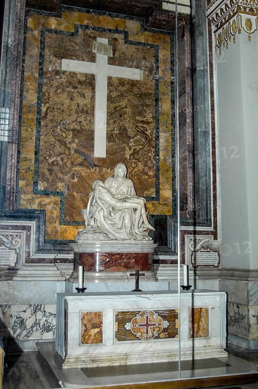 View of niche small side aisle in title Basilica maior San Pietro Papal Basilica St Peter's Basilica with marble sculpture La Pieta by Michelangelo Buonarotti High Renaissance sculptor with Mary Mother of God Mother of God Holy Virgin holding crucified Jesus Christ Saviour Son of God in her arms, behind large cross with inscription plaque Inri, St Peter's Basilica, Vatican City, Rome, Lazio, Italy