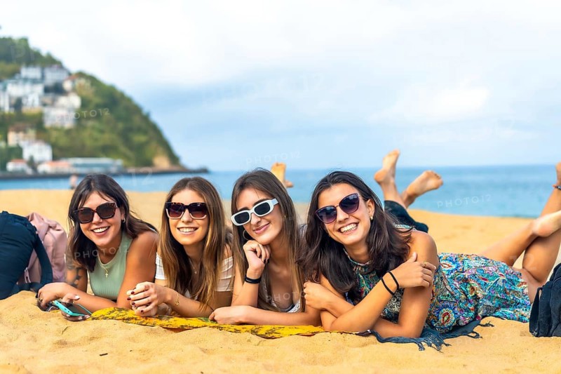Four happy young women friends lying on a towel enjoying summer vacation on a sandy beach, having fun and smiling at the camera with the ocean and a coastal town in the background