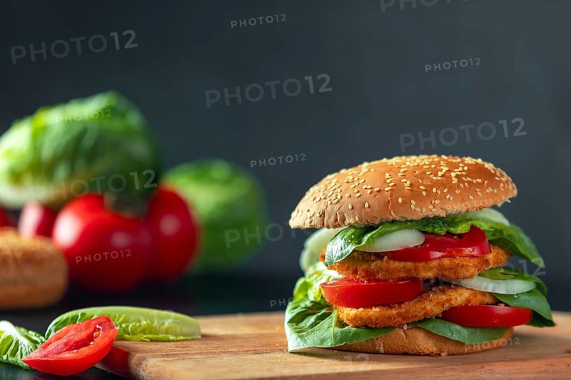 Burger with schnitzel, tomatoes, onions and lettuce on a wooden board, ingredients in the background