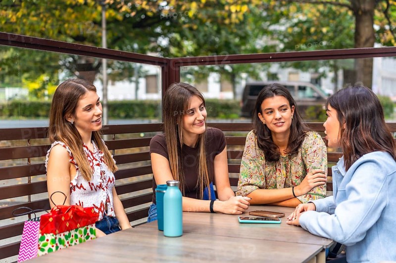 Four young women chatting and laughing at an outdoor cafe table after a shopping spree, enjoying coffee, gift bags and relaxed city side socializing on a sunny daytime outing