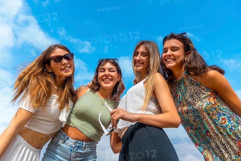 Group of young women standing close together outdoors, smiling and laughing on a sunny summer day under a bright blue sky, enjoying friendship, freedom and carefree joy