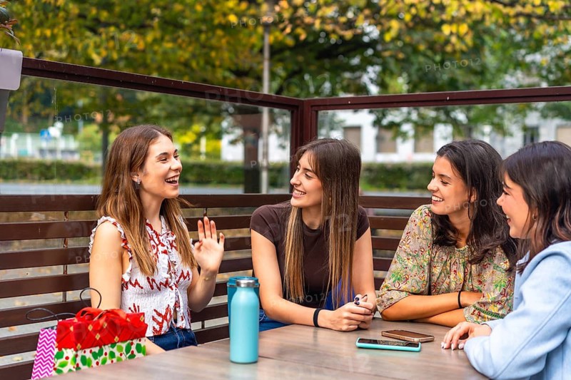 Four young women friends enjoying conversation and shared laughter at a cafe table outdoors, holding shopping bags and relaxing together after a day of leisure activities
