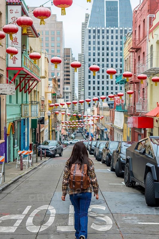 Young person with a backpack walking up a vibrant san francisco chinatown street adorned with traditional red lanterns, passing historic buildings and parked cars