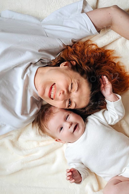 A joyful mother and her adorable baby girl lying together on a cozy bed, both dressed in white. The scene exudes warmth, happiness, and family bonding