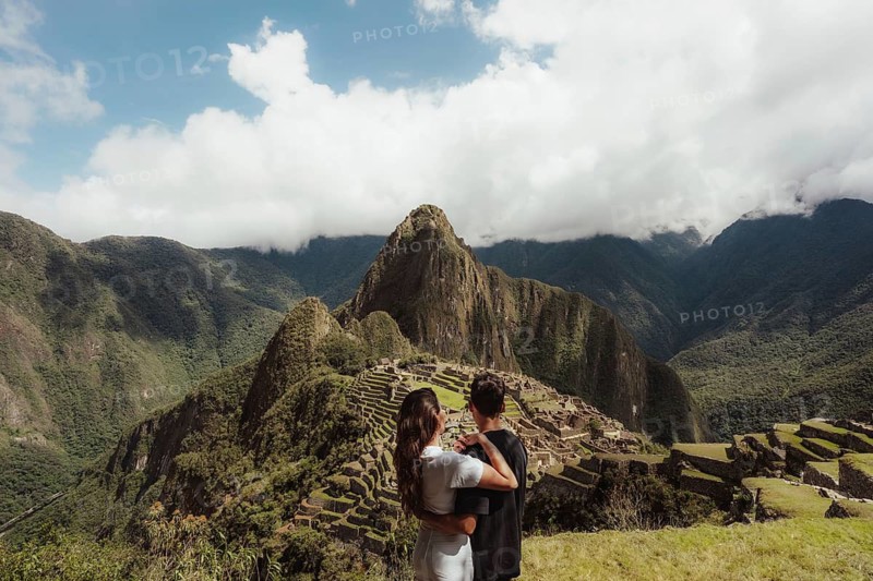 A couple stands atop a hill in Peru, gazing at the majestic ruins of Machu Picchu surrounded by lush green mountains and a cloudy sky, capturing a moment of wonder