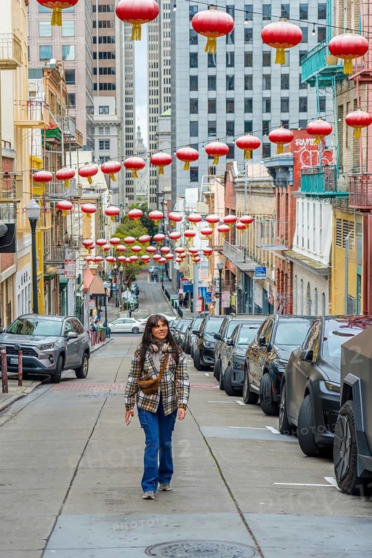 Young woman confidently walking along a street in san francisco's chinatown, surrounded by traditional red lanterns above, parked cars, and historic buildings in an urban setting