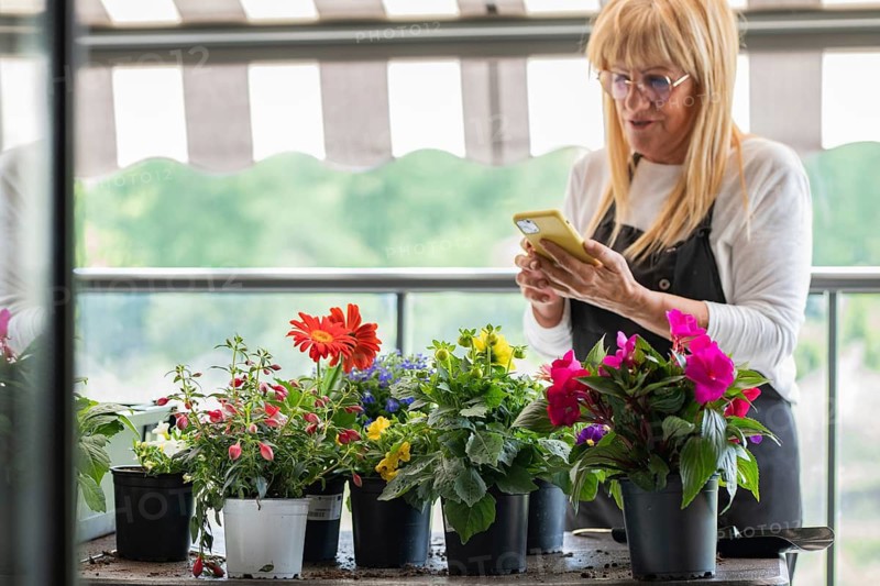 A woman stands on her balcony tending to vibrant potted flowers, capturing the moment on her smartphone. The scene reflects a blend of gardening, nature and technology