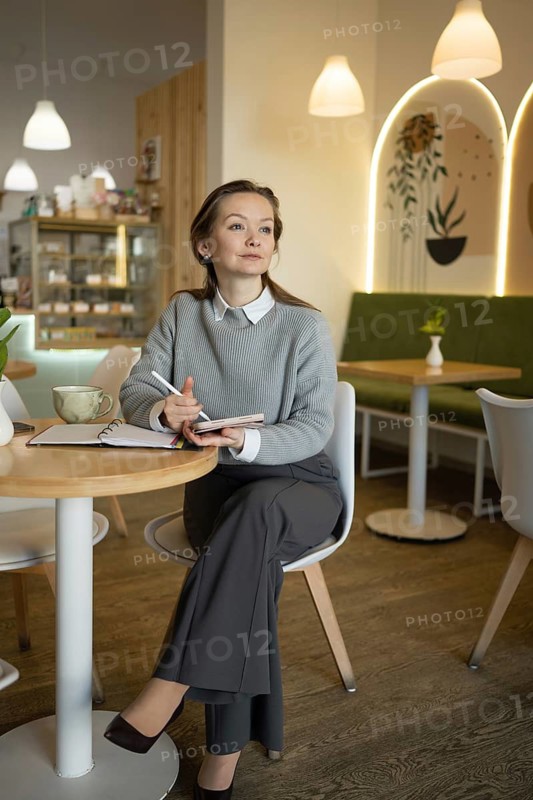 A woman in a stylish outfit sits thoughtfully in a cozy cafe, holding a pen and notebook. The warm ambiance is enhanced by soft lighting and contemporary decor