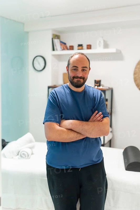 Professional male masseur standing with crossed arms, smiling at the camera in a modern, serene spa room, ready to provide soothing wellness and therapeutic body treatments
