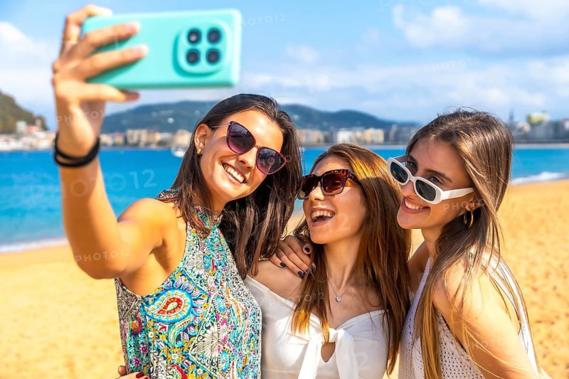 Three smiling young women friends taking a selfie with a smartphone, enjoying their summer vacation at the beach with a vibrant blue sea and sunny sky in the background