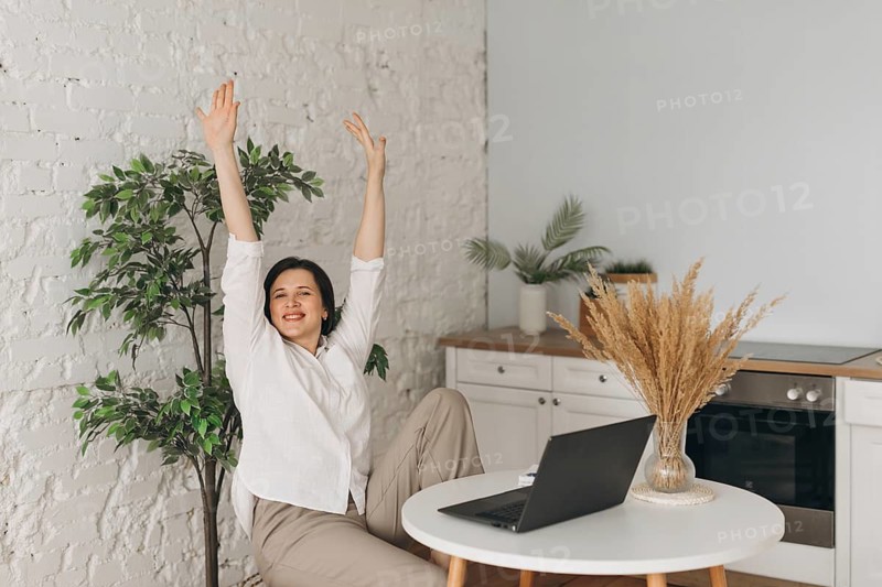 A cheerful nutritionist takes a rejuvenating break in her modern kitchen office. With a laptop on the table, she stretches, surrounded by greenery and minimalist decor