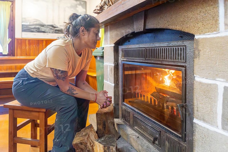 A woman warms herself by a cozy fireplace in a rustic shelter in Gorbea Natural Park, Basque Country, Spain. The serene setting offers a peaceful retreat in nature