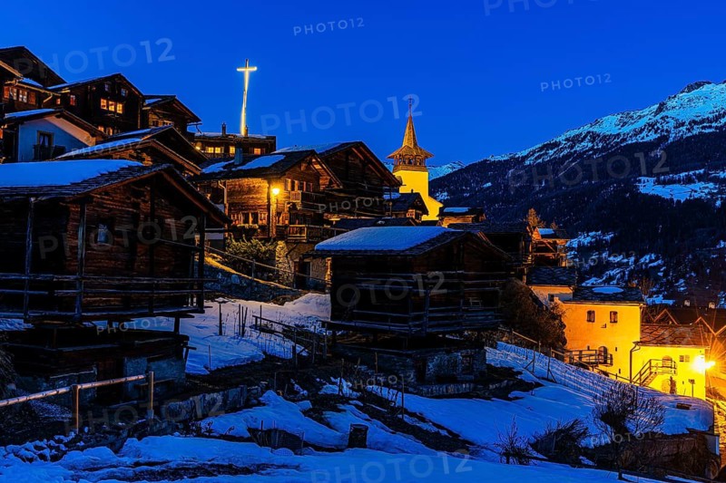 Snow-covered roofs of old farmhouses and barn storage, in the back the illuminated cross and the illuminated church tower of the Saint Theodule church, night view, historic town center, Grimentz, Val d'Anniviers, Valais Alps, Canton of Valais, Switzerland