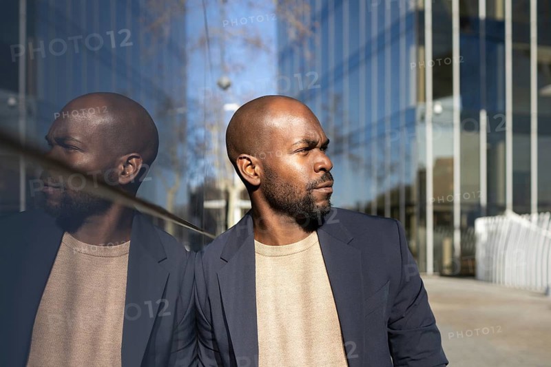 Confident cuban man in a blazer stands near a reflective glass wall, his reflection visible. Modern architecture and blue sky create a professional atmosphere