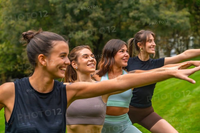 Four happy young women practicing warrior pose during an outdoor yoga session, promoting wellness, friendship, and a healthy lifestyle in a green natural environment