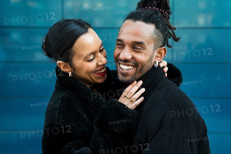 A happy multicultural couple embraces and smiles lovingly at each other, symbolizing joy and connection. They are wearing black coats against a modern blue background