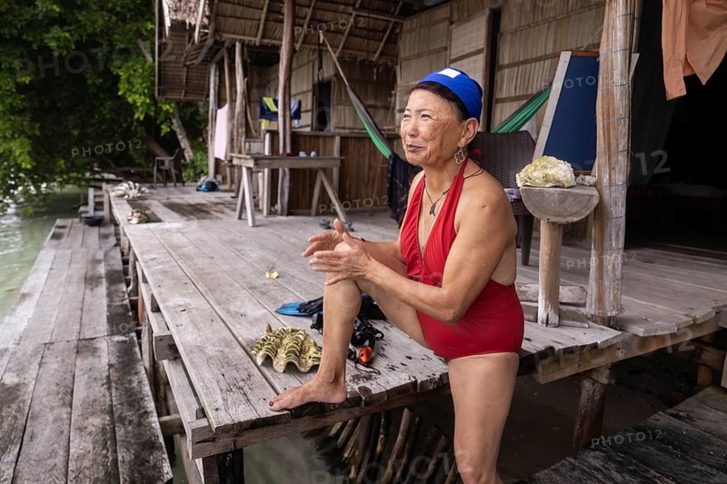 An elderly Asian woman sits cheerfully in a red swimsuit on a rustic wooden jetty in Kri Island, Raja Ampat, Indonesia, surrounded by lush greenery and waterfront scenery