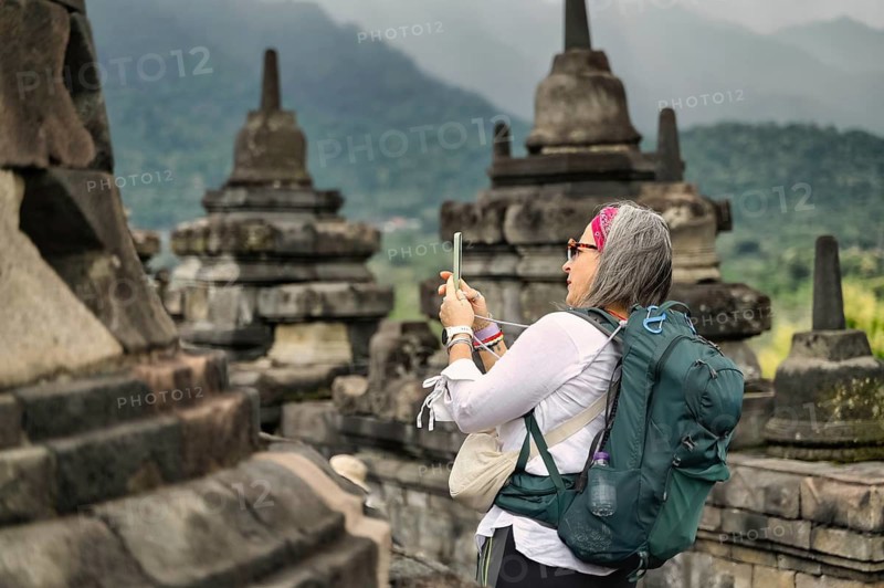 Side view of a female senior tourist capturing photos with a smartphone at the historic Borobudur Temple, framed by ancient stone stupas against a backdrop of misty mountains