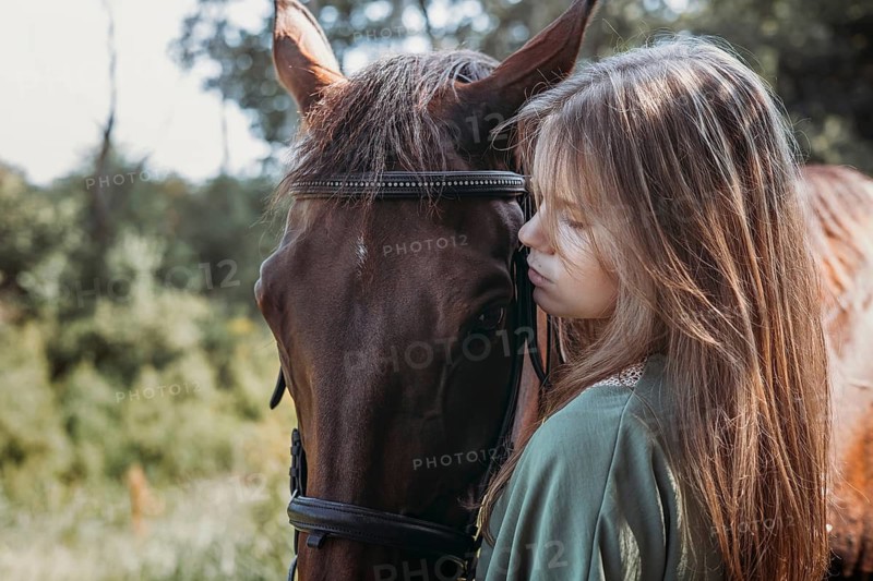 A girl with long hair gently embraces a brown horse in a sunlit forest. The scene captures the serene connection between human and animal amidst nature