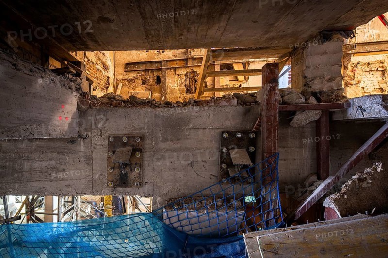 A dynamic construction site scene features exposed concrete, steel beams, and protective netting. Sunlight highlights the rugged textures and ongoing transformation