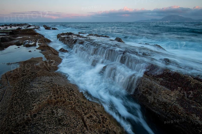 The rocky coast of Tarifa, Cadiz, during sunset. Waves gently cascade over rocks, capturing the serene beauty of the Atlantic coastline under a colorful sky