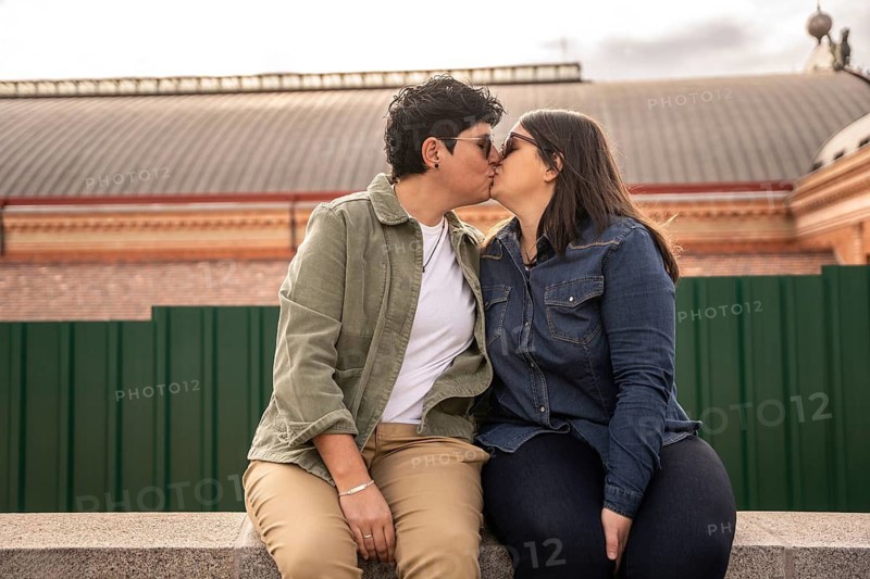 A curvy lesbian couple in casual attire shares a heartfelt kiss outdoors, symbolizing love and connection. The intimate moment captures diverse representation and affection
