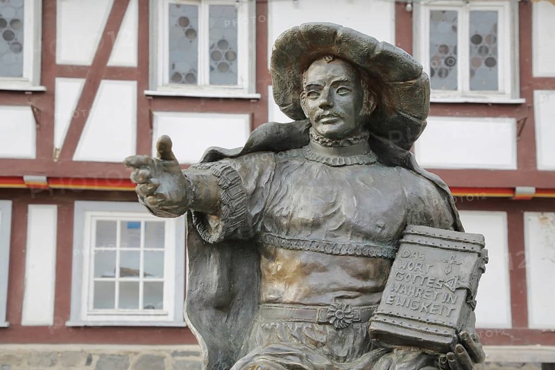 Philip the Magnanimous sculpture by Ewald Rumpf 2001, modern art, bronze sculpture, life-size, sitting man with historical clothing and Bible with inscription, The word of God would be eternal, Hessian church history during the Reformation, gesture, arms, index fingers, Philip I of Hesse, marketplace, Homberg-Efze, Hesse, Germany