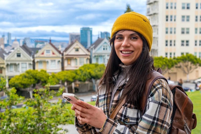 Young woman smiling at the camera, holding a smartphone, with the iconic colorful victorian houses known as the painted ladies and the san francisco skyline visible in the blurred background
