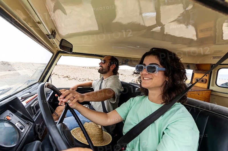 A blind man sits in the passenger seat while a woman drives a motorhome through a scenic desert. Their smiles highlight adventure, teamwork, and the joy of exploration