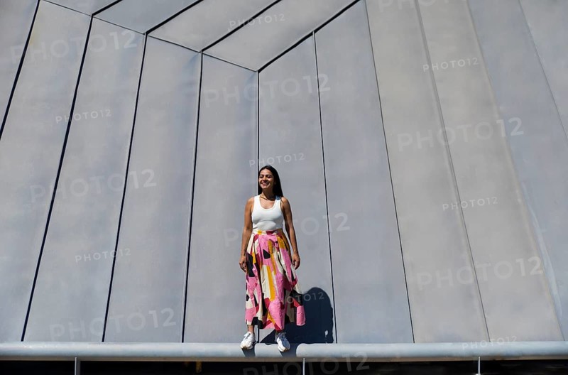 A woman in a vibrant, colorful skirt stands confidently in front of a striking piece of modern architecture. The contrast between her outfit and the urban setting is captivating