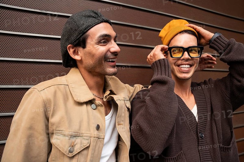 A cheerful couple stands side by side against a modern metal wall. Both are dressed in casual urban attire, with one wearing a beanie and glasses, the other a cap. They appear relaxed and happy