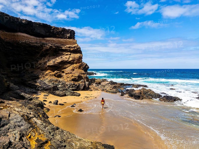 From above a lone woman walks on the sandy shore of Tindaya Beach in Fuerteventura, amidst rugged cliffs and foamy sea waves