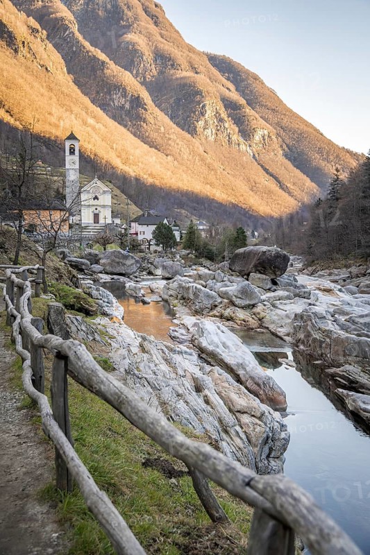 A picturesque view of Lavertezzo, Switzerland, featuring a charming church, rustic wooden fence, rocky riverbed, and majestic mountains bathed in golden sunlight during late afternoon