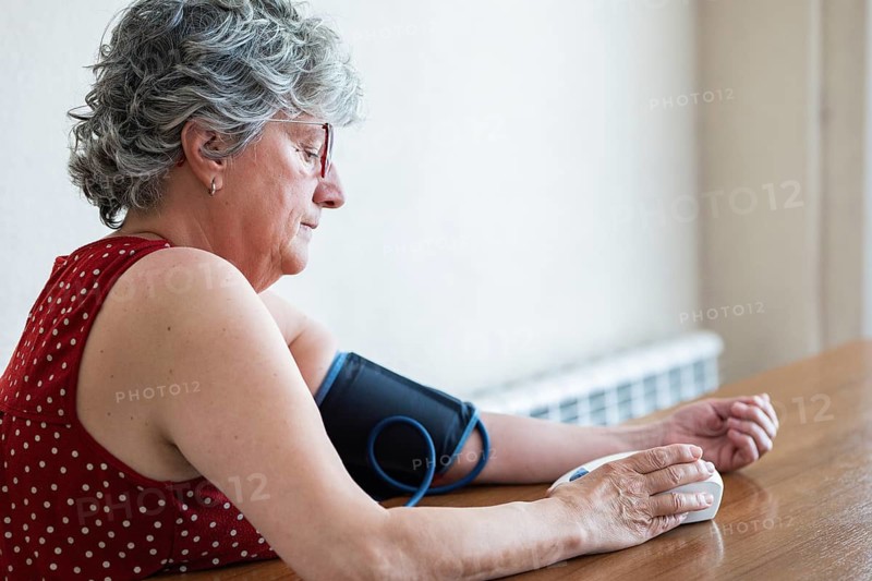 An elderly woman is measuring her blood pressure at home using a digital monitor. She appears calm and focused, highlighting the importance of health awareness and self care