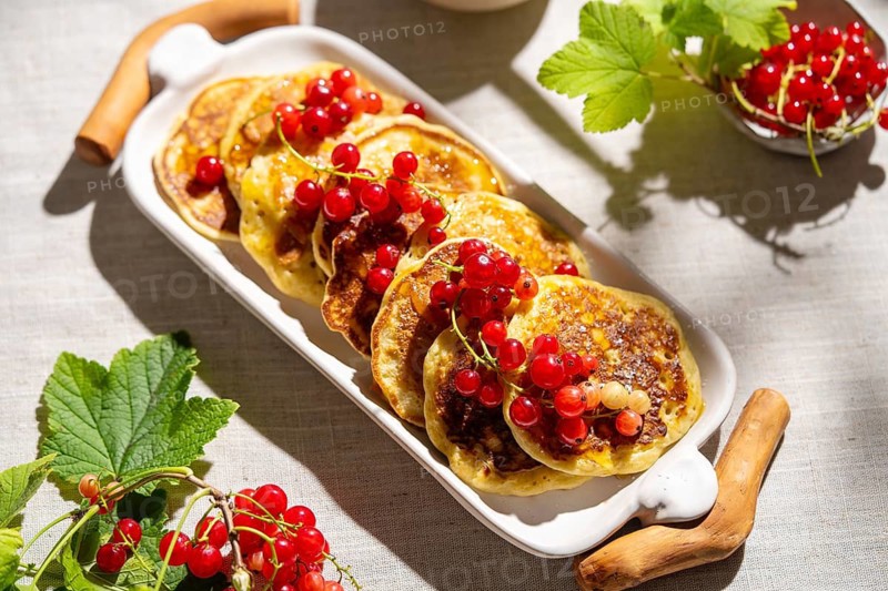 Top view of fluffy pancakes topped with vibrant red currants, elegantly arranged on a white ceramic plate. The light beige linen fabric adds a soft, rustic charm