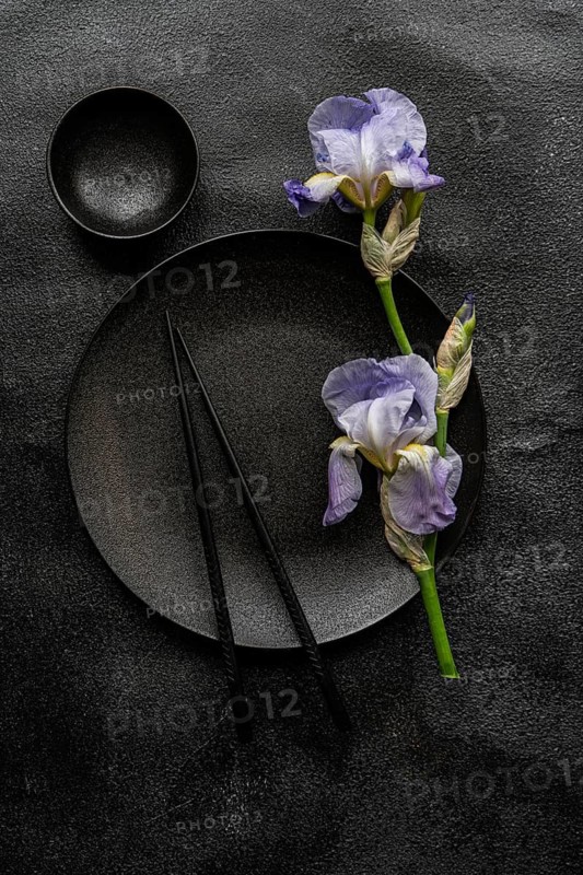 Top view of elegant iris flowers alongside black ceramic dishware on a textured dark background, creating a striking visual contrast that emphasizes simplicity and natural beauty in table setting decor