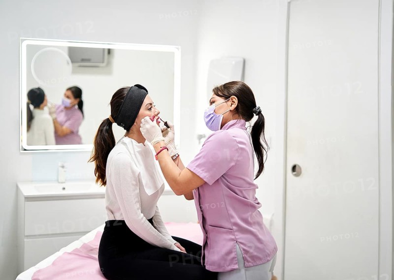A beauty therapist in a lavender uniform and mask carefully applies lip color to a seated client wearing a headband and bib, in a clean and modern aesthetic clinic environment