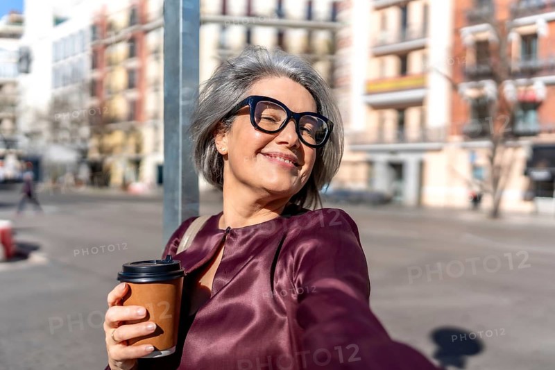Mature woman with gray hair and glasses smiling for a selfie on a sunny city street, holding a takeaway coffee cup, enjoying a carefree, confident moment outdoors