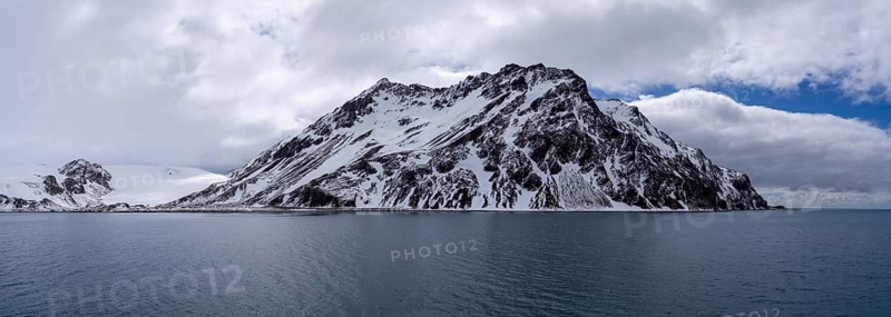 Snow-capped mountain with rugged rock formations rising above the ocean, cloudy sky, coastal landscape, South Georgia, South Atlantic