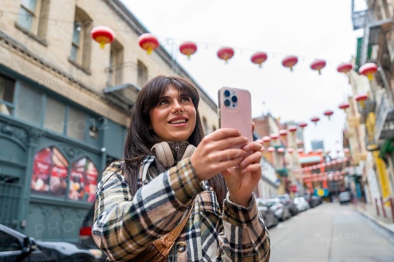 Woman standing on a san francisco chinatown street, looking up and smiling while capturing the vibrant red lanterns and historic architecture with her smartphone, experiencing travel and tourism