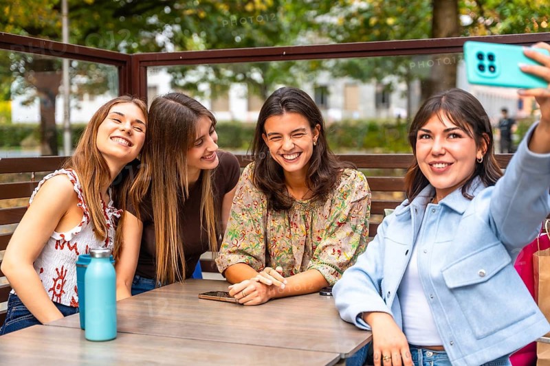 Group of young multiethnic women sitting at an outdoor cafe table, smiling and laughing as they take a selfie with a smartphone, enjoying a casual social moment together