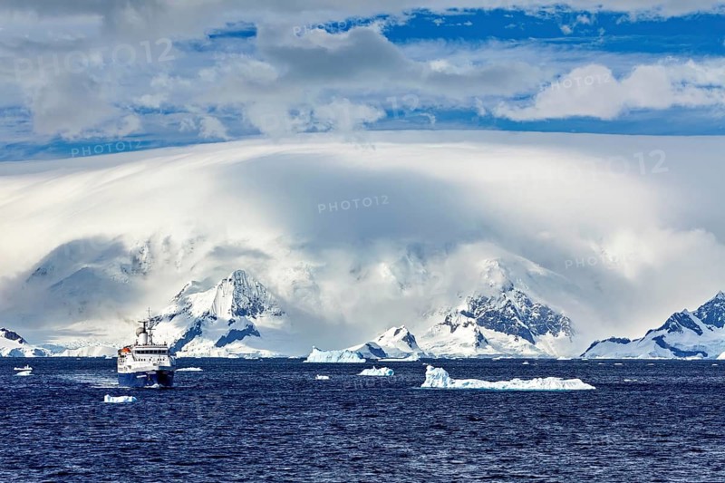 A ship sails in the ocean, surrounded by icebergs and snowy mountains under a cloudy sky, cruise ship in the Southern Ocean in Antarctica