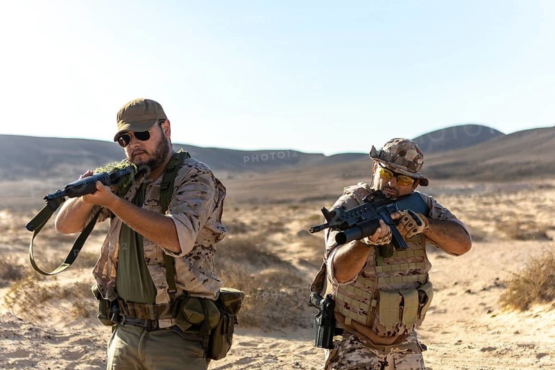 Two armed soldiers dressed in camouflaged tactical gear and sunglasses, aim rifles in a desert environment, portraying military readiness and strategic mission operation