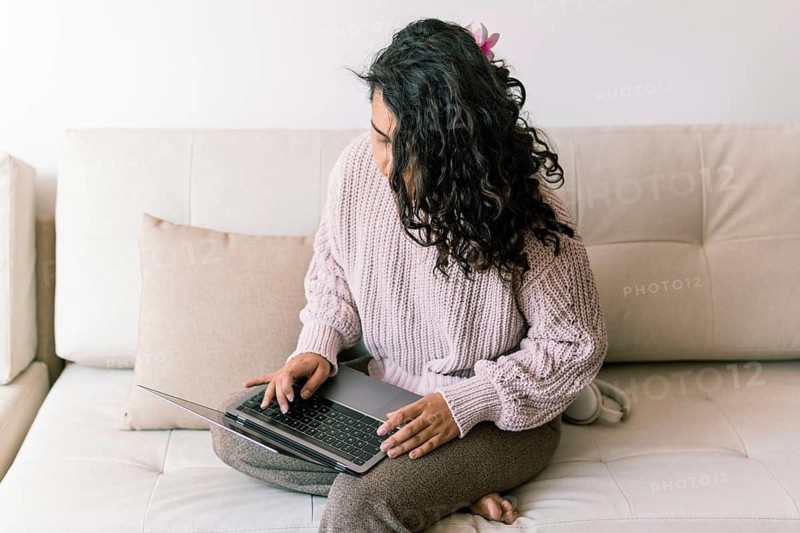 A young woman sits comfortably on a sofa at home, using a laptop She wears a cozy sweater and seems engaged in her task, surrounded by a calm and inviting atmosphere