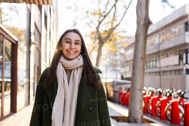 A cheerful woman in a green coat smiles warmly on a sunny day in an urban environment She is wearing glasses and a light scarf, with blurred city buildings and vehicular traffic in the background
