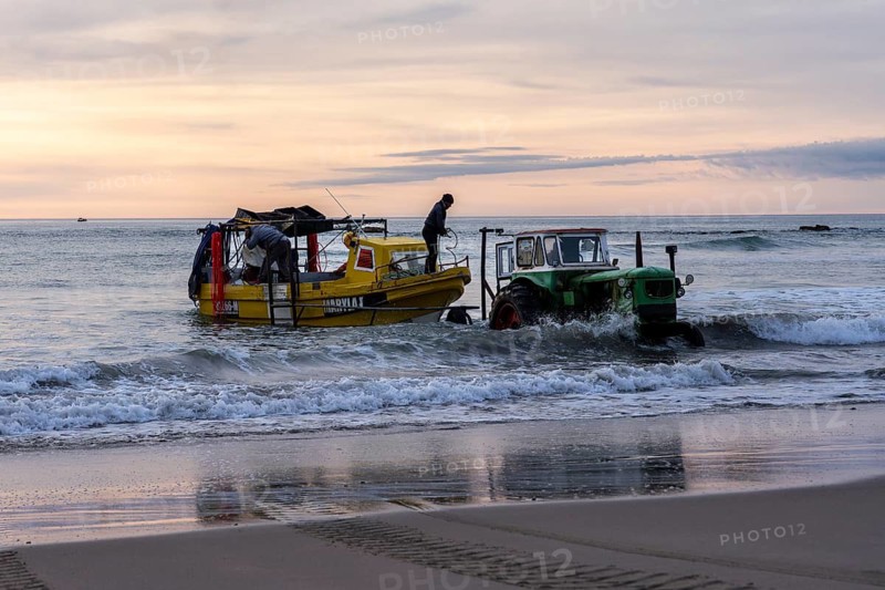 Yellow fishing boat with two fishermen is pulled from beach into water by green tractor at sunrise, Playa Union, Rawson, Chubut Province, Argentina