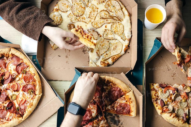 Top view of a group of unrecognizable friends enjoying assorted pizzas together. Various hands, both male and female, reach into cardboard boxes to grab slices, showcasing a casual dining experience
